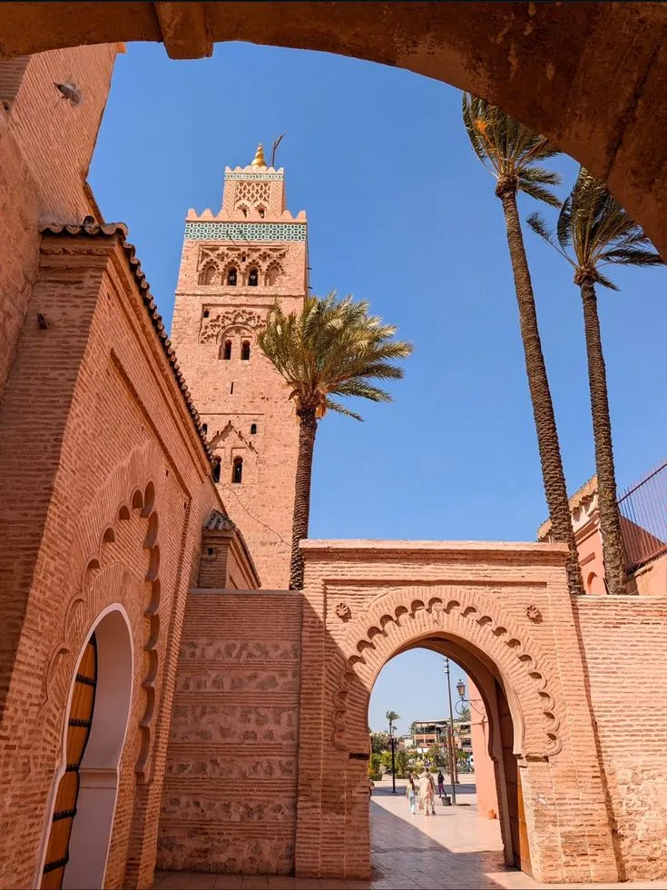 Minaret de la Koutoubia à Marrakech vu à travers une arche en brique rouge, entre les palmiers
